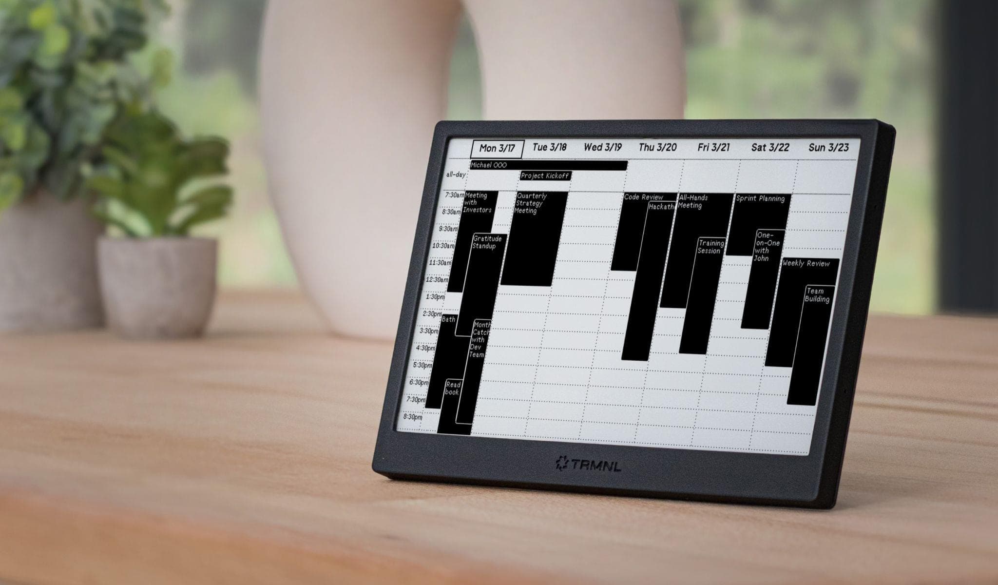 TRMNL is a small, black tablet-like desktop display on a light wooden tabletop showing a weekly calendar from Mon 3/17 to Sun 3/23 with an all-day row and multiple filled time slots listing work items such as “Project Kickoff,” “Code Review,” “All-Hands Meeting,” and other tasks. The scene depicts a calm home-office workspace with a softly blurred background including a leafy plant and a large white vase.