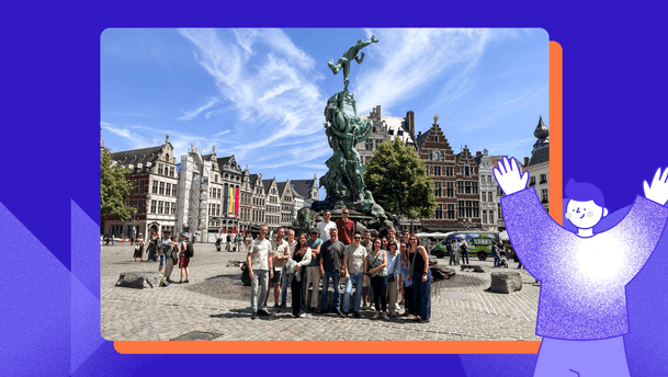 Brabo Fountain, Antwerp – a group of around twenty adults pose for a sunny city-centre group photo on Grote Markt, with the Brabo Fountain rising behind them amid ornate Flemish guild houses and the City Hall. The image depicts a bright day with a vivid blue sky and a cheering illustrated character on the right.